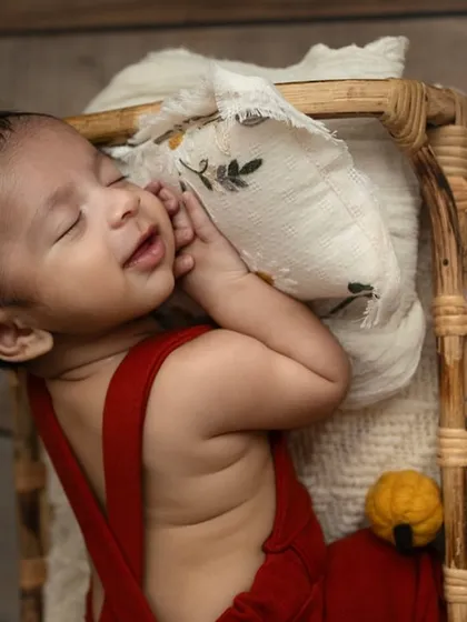 A sweet newborn moment, with the baby smiling in their sleep while resting in a tiny wicker bed. The pop of red from the overalls adds a playful touch.