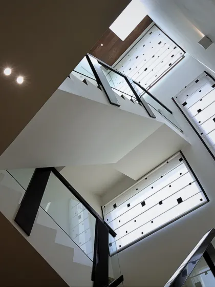 An upward view of a staircase, showcasing the clean geometry of the glass and black metal railings against uniquely patterned window blinds that play with light and shadow.