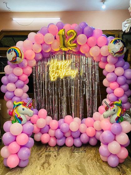 A unicorn-themed balloon arch for a 12th birthday, featuring a silver fringe backdrop, unicorn head balloons, and a neon sign.