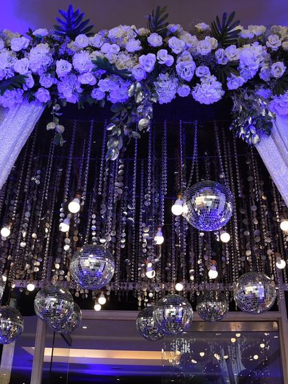 A close-up of a glamorous entrance detail. A canopy of white flowers is accented with hanging disco balls and Edison bulbs, blending romantic florals with a fun, party-ready vibe.