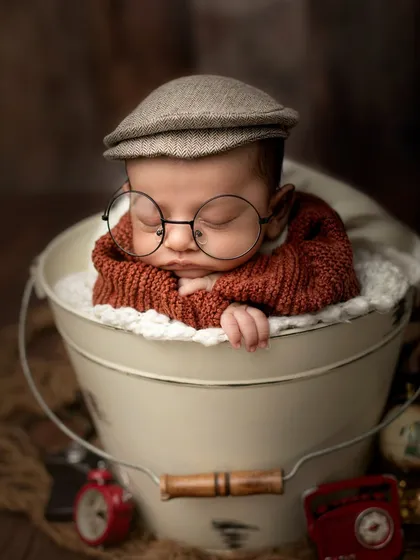 A vintage-inspired portrait of a baby boy in a bucket, complete with a newsboy cap and oversized glasses for a touch of old-world charm.