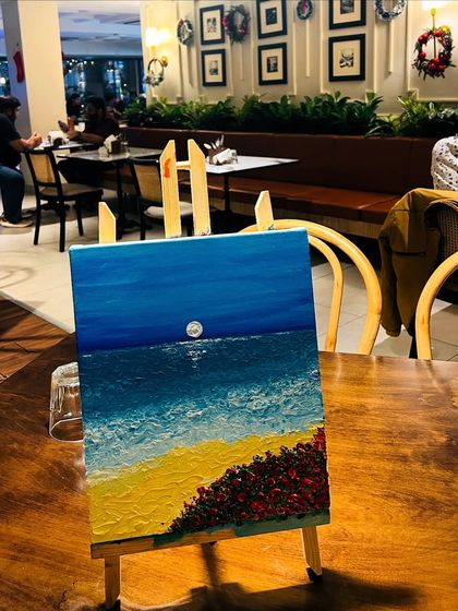A finished knife painting of a beach scene, resting on an easel in a cozy cafe setting, waiting to be taken home.