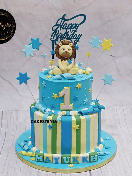 A cheerful two-tier jungle theme cake for a first birthday. This 3kg choco-vanilla cake features a cute lion topper and a striped bottom tier with the child's name in fondant blocks.