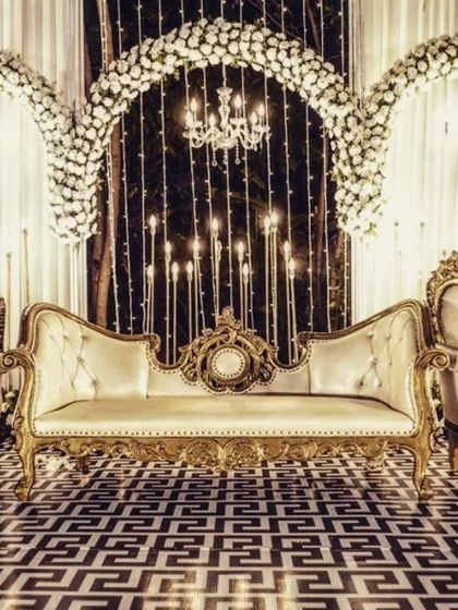 A classic and opulent reception stage with ornate gold and cream furniture, set against a backdrop of white floral arches and twinkling lights.