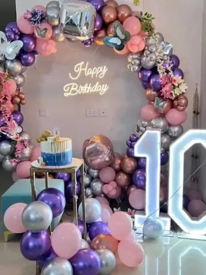 A 10th birthday party with a circular balloon arch in purple, pink, and silver. The setup includes a "Happy Birthday" neon sign, a cake stand, and a marquee number '10'.