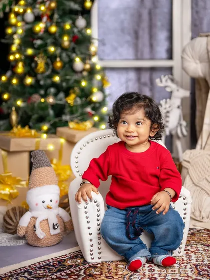 A happy baby boy in a bright red sweater, sitting contentedly in his chair amidst the festive Christmas decorations.