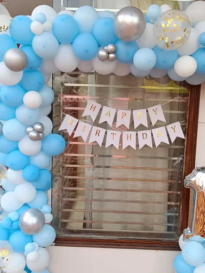 A simple and classic first birthday balloon decoration for a home celebration. This L-shaped arch in blue, white, and silver frames a window, complemented by a 'Happy Birthday' banner and a large number '1' foil balloon.