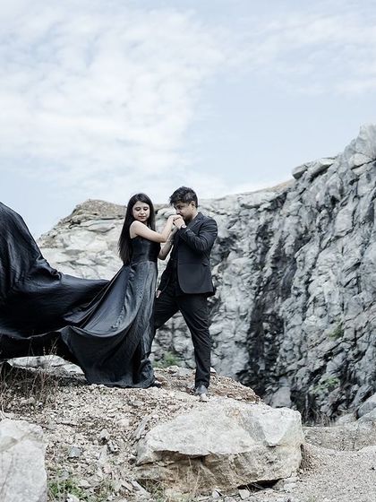 A tender moment where the groom kisses the bride's hand, their formal black attire contrasting with the rugged natural location and the flowing gown.