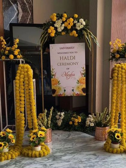 A welcoming entrance for a Haldi or Megha ceremony. The setup includes a personalized welcome sign, floral arrangements with marigolds, and decorative stands.
