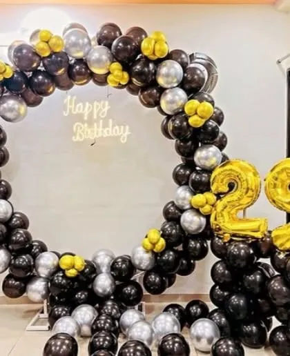 A 29th birthday party with a circular balloon arch in black, silver, and yellow. The setup includes a "Happy Birthday" neon sign and foil number balloons.
