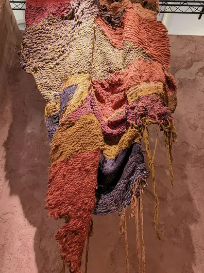 A close-up of a richly textured, multi-colored hanging textile from the 'Soil' exhibition. The piece uses thick, hand-spun yarns to create a surface that resembles a cross-section of earth.