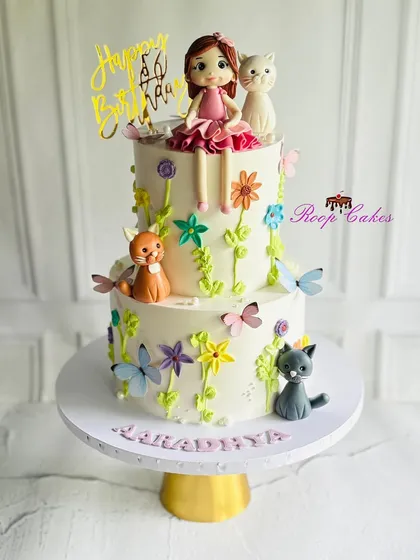 Whiskers, flowers, and birthday magic. This adorable two-tier cake is bursting with charm, featuring playful cats, blooming flowers, butterflies, and a sweet little doll topper.