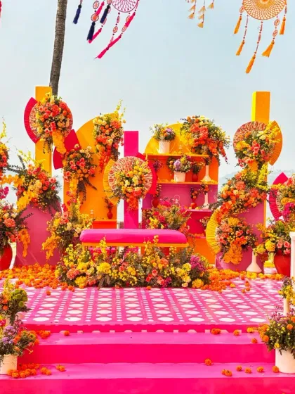 A vibrant and modern outdoor stage for a Haldi or Mehndi ceremony, using a bold color palette of pink and yellow, with abundant marigold and rose floral arrangements.