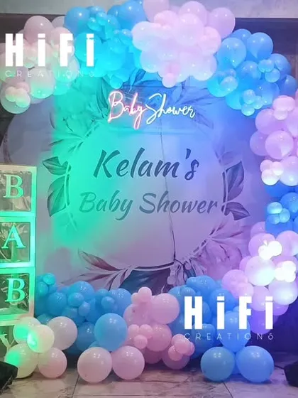 A blue-themed baby shower for welcoming a baby boy. The decor features a balloon arch, "BABY" boxes, and character cutouts, all illuminated with cool lighting.
