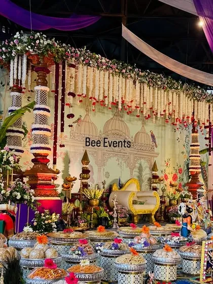 A wide view of a luxurious and traditional decor setup for a Half Saree ceremony. The scene is filled with intricate details, from the floral 'thomala' pillars to the peacock motifs and the elaborate display of sweets and gifts.