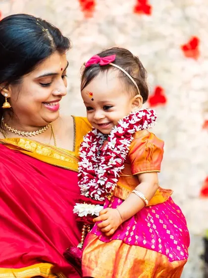 A tender moment between a mother and her baby during a traditional Ayusha Homam ceremony, with the baby wearing a flower garland.