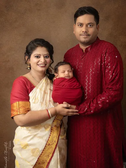 A classic family portrait in traditional Indian attire. The parents, dressed in red and white, hold their newborn, creating a culturally rich and beautiful family memory.