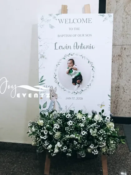 This charming welcome board was designed for a baby boy's baptism. It features a cute rabbit illustration and a photo of the child, complemented by a base of fresh baby's breath and white flowers.