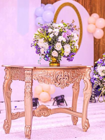 An exquisite floral arrangement on an ornate golden table. The purple and white flowers perfectly complement the royal theme of the event.