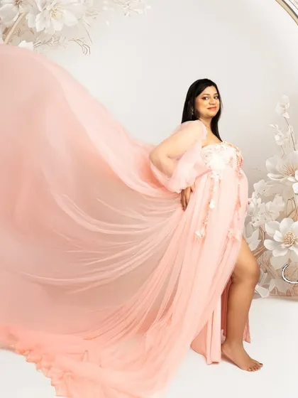 A light and airy fine art portrait. The expecting mother poses in a soft pink gown with a long, flowing train against a clean white backdrop, creating a feeling of grace and elegance.