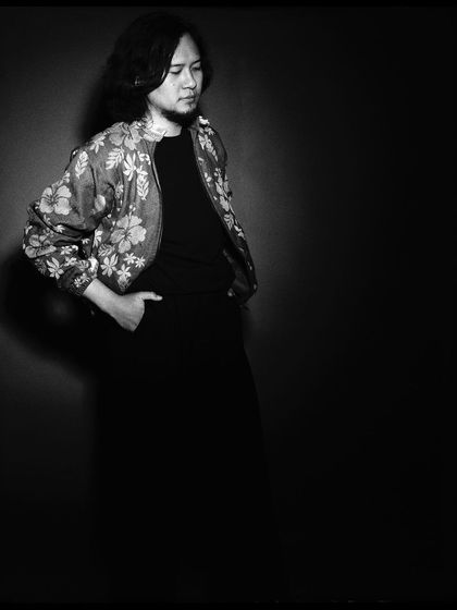 A moody, low-key black and white portrait of Taba Chake. The focus is entirely on his form and the texture of his jacket.