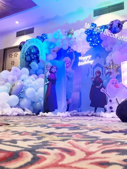 A wide view of a Frozen-themed stage decoration for a 5th birthday. The setup is filled with balloon clouds, character cutouts, and a large illuminated number '5', creating an immersive experience.