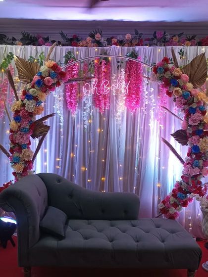 A vibrant and colorful floral arch decoration for a baby shower. This setup includes a 'Oh Baby' neon sign, hanging wisteria flowers, and a comfortable chaise lounge for the guest of honor.