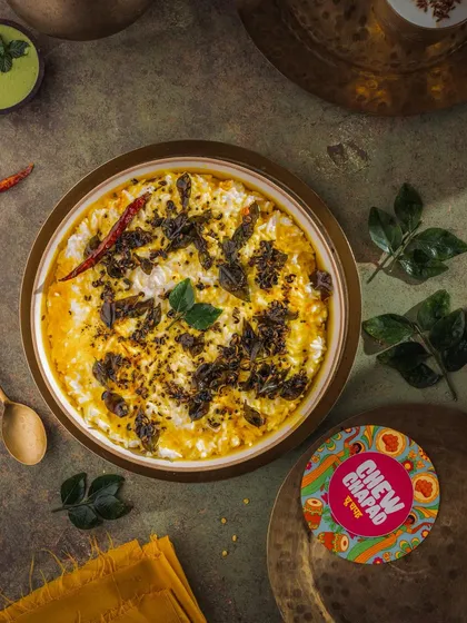 A top-down view of tempered curd rice, styled for Chew Chapad. The dark, moody lighting makes the vibrant yellow of the tempering and the fresh curry leaves pop, emphasizing the dish's comforting yet flavorful nature.