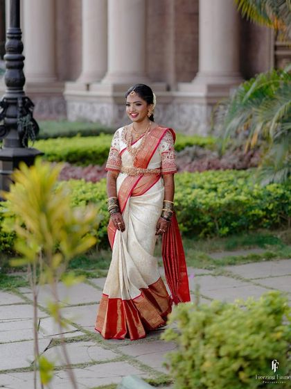 A bride in a stunning white and red Kanchipuram saree, walking through a palace garden.
