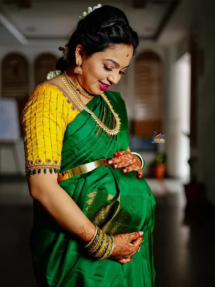 A solo portrait of the mother-to-be, looking down at her bump with a gentle smile. The lighting is set to accentuate the rich colors of her saree and the happy glow on her face.