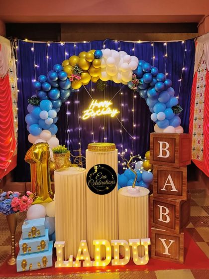 A blue and gold themed first birthday party at home. The decor includes a balloon arch over a blue curtain with fairy lights, creating a magical effect.