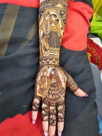 This bridal design features two distinct couple portraits. The palm shows a wedding ceremony, while the forearm depicts Radha and Krishna, blending personal and spiritual love stories.