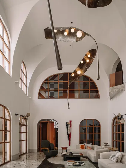 A wide shot of the double-height living room at Rehethaan, showcasing the grotto-like architecture, arched windows, and the magnificent leaf-like light installation that hangs from the ceiling.