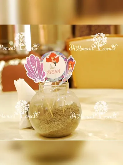 A small jar filled with sand and decorated with mermaid-themed toppers serves as a creative and unique table detail or party favor.