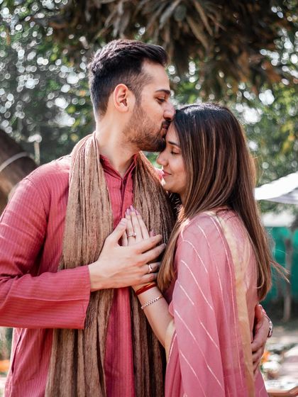 A tender moment where he kisses her forehead as they hold hands. The outdoor setting and their traditional attire create a beautiful blend of culture and nature.