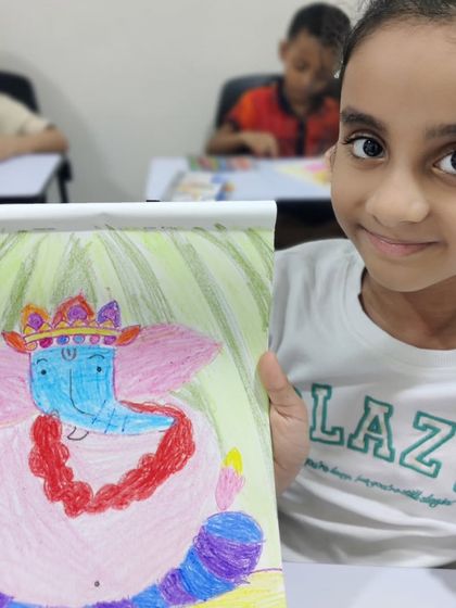 A young girl smiles as she presents her Ganesha drawing. She gave her Ganesha large pink ears and a beautiful red garland, showcasing her unique artistic choices in this guided drawing session.
