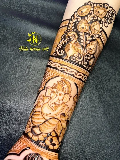 A close up of the arm portion of a bridal design, showing a detailed figure of Lord Ganesha. Above it is a beautiful peacock feather band, combining two auspicious symbols.