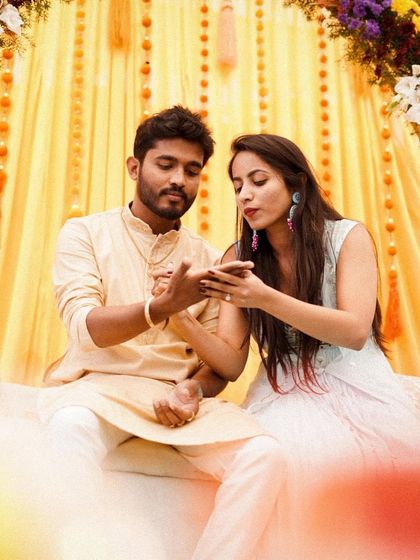A candid moment of the couple admiring the henna design. The warm yellow backdrop with hanging tassels adds to the festive and celebratory mood of the Mehendi function.