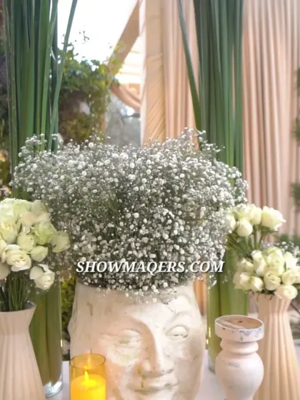 A decor detail featuring a unique planter with a face sculpture, filled with baby's breath and surrounded by white roses and candles.