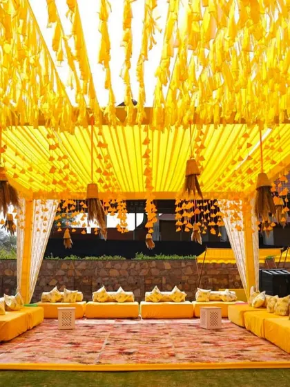 A cozy and traditional Haldi seating arrangement under a canopy decorated with yellow tassels and drapes. The low seating with printed cushions creates a comfortable and intimate atmosphere for guests.