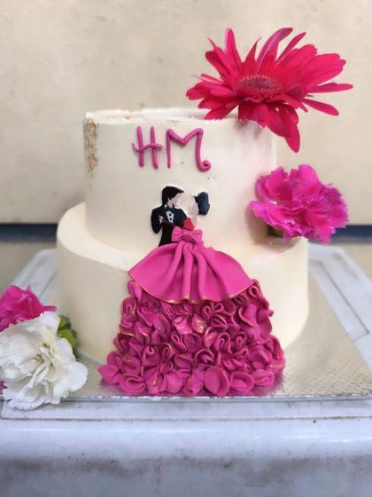 A vibrant anniversary cake that's full of life. The highlight is the hand-piped pink ruffle dress on the edible couple cutout, complemented by bright fresh gerbera daisies.