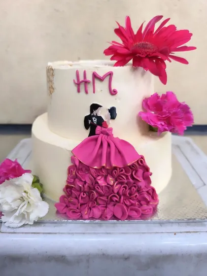 A vibrant anniversary cake that's full of life. The highlight is the hand-piped pink ruffle dress on the edible couple cutout, complemented by bright fresh gerbera daisies.