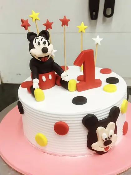 A classic Mickey Mouse cake for a first birthday celebration. It features a large fondant Mickey figurine, star toppers, and the iconic polka dot pattern.