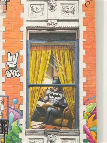 In this scene from "Ordinary Powers," Batman is caught in a quiet moment, reading a book behind a window adorned with NYC graffiti.