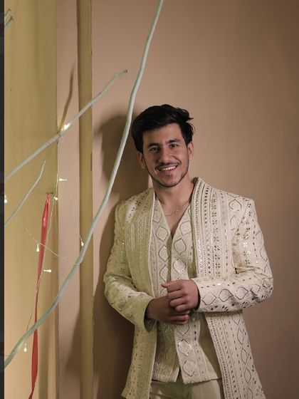 A client smiling in our off-white Indo-Western jacket set. The outfit features a long jacket with geometric mirror work, worn over a matching waistcoat and trousers.