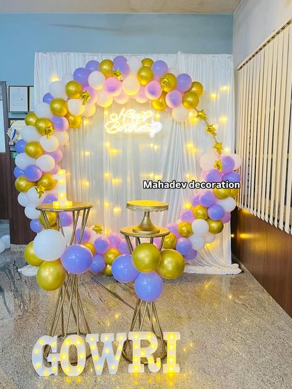 A beautiful and simple birthday setup with a circular frame decorated with pastel purple, gold, and white balloons. The white curtain backdrop with fairy lights creates a soft, dreamy effect, perfect for photos.