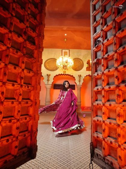 A dramatic entrance. The bride-to-be twirls in her magnificent magenta lehenga, framed by grand wooden doors, creating a powerful and regal portrait.
