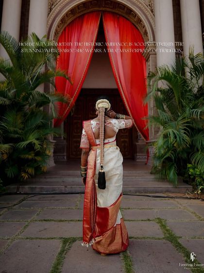 The back of the bride in her traditional white and red saree, showcasing her intricate hairstyle as she walks towards a grand entrance.