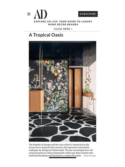 A magazine feature showcasing a tropical-inspired bar area. The dark, moody wallpaper with a vibrant botanical print creates a stunning focal point, complemented by a sleek wooden bar and crazy-paved black marble flooring.