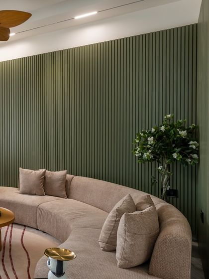 The curved sofa's soft form contrasts beautifully with the rigid vertical lines of the olive green fluted wall. A wooden fan adds a touch of natural warmth to the ceiling.